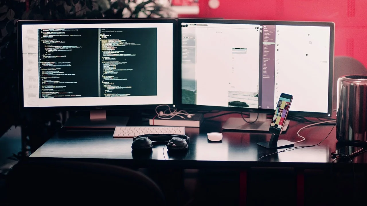 desk setup with two monitors, keyboard and a pair of headphones with a smartphone on a stand on the right-hand side 