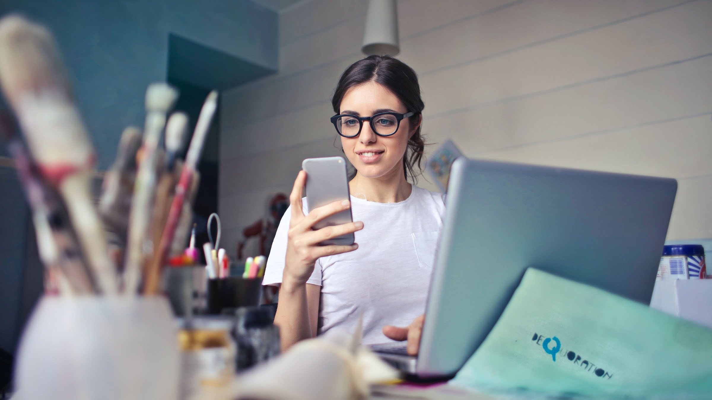 woman using a smartphone with a laptop and documents on the table in front of her
